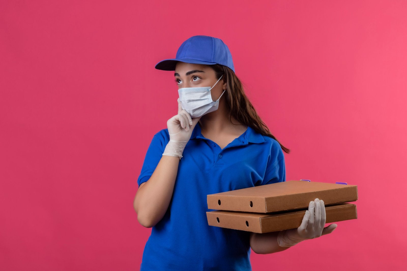 young delivery girl in blue uniform and cap wearing facial protective mask and gloves holding pizza boxes looking aside with pensive expression thinking standing over pink background